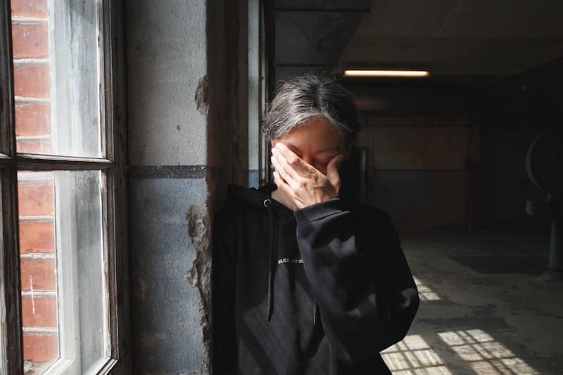 Person covers face with hand near window.