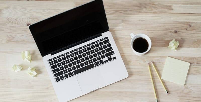 Image of a laptop, a cup of coffee, a notepad, and pencil on a clear wooden desk.