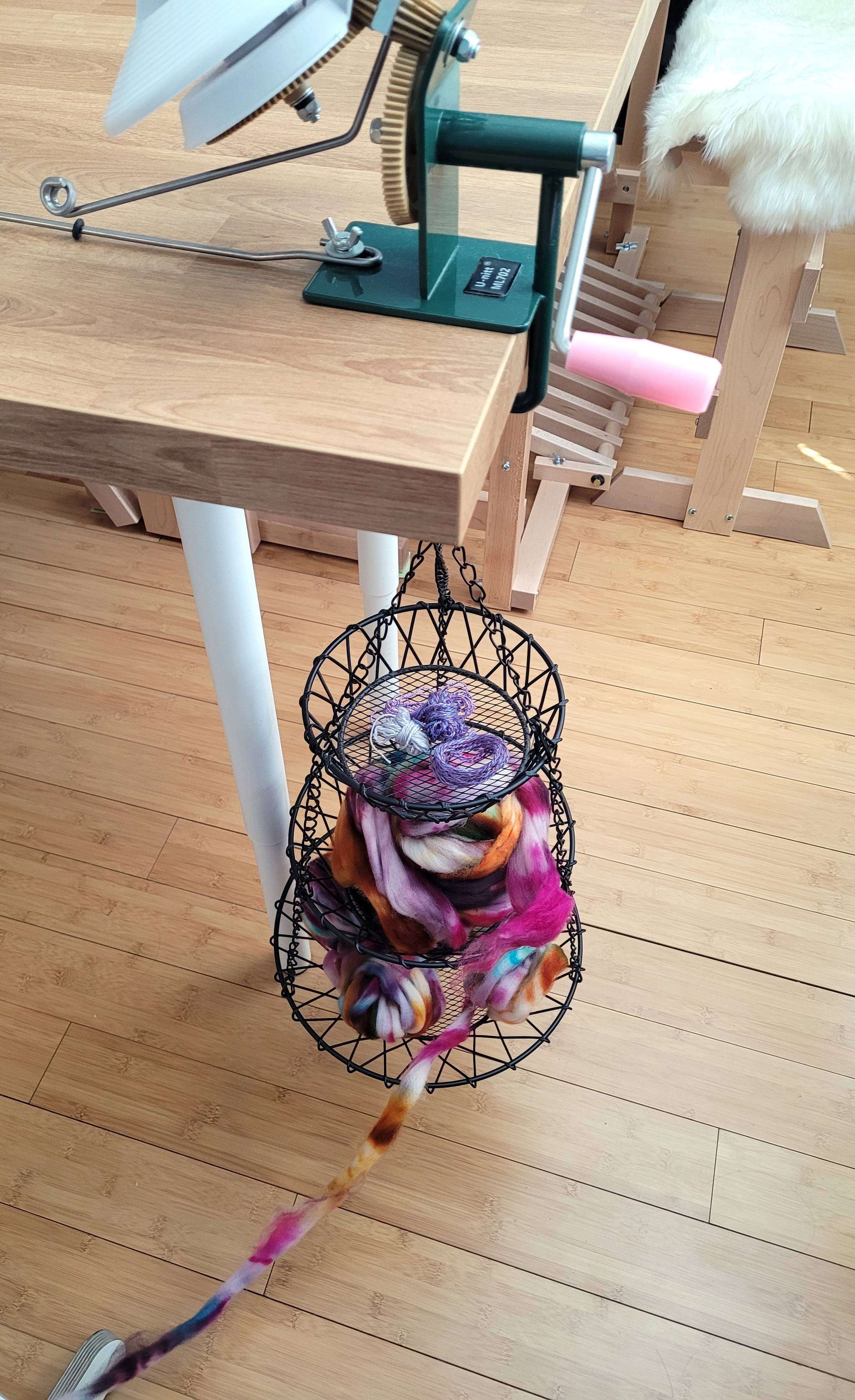 A black, wire, three-tier basket hangs from the end of a wooden work table with a yarn winder mounted above. The three-tier basket is filled with multicolored fiber on the two lower tiers, and some leftover handspun yarn bits on the uppermost tier. A loom bench and treadles can be seen in the background.