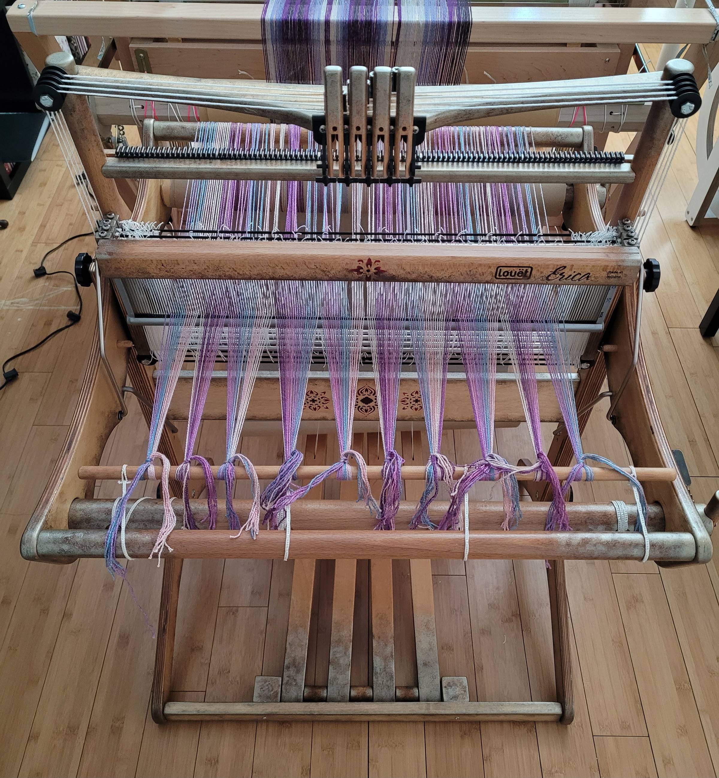 Louet Erica four-shaft table loom (top view), positioned on its stand, and fitted out with a set of four treadles. There is a purple/pink/blue striped warp on the loom, and it's ready to start weaving. The loom itself has a custom, weathered-looking, metallic paint job around the edges, and some floral stencils painted on.