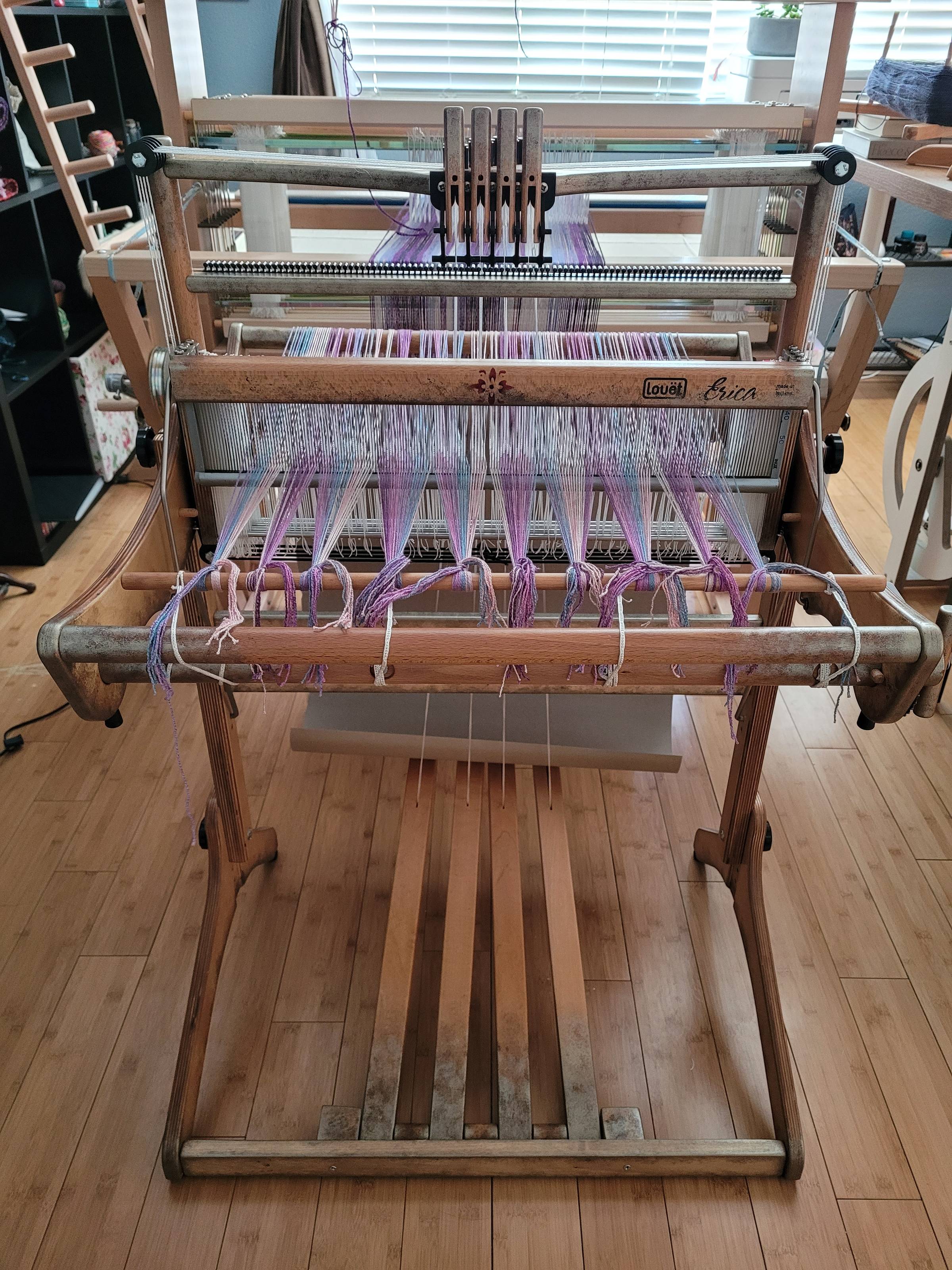Louet Erica four-shaft table loom (front view), positioned on its stand, and fitted out with a set of four treadles. There is a purple/pink/blue striped warp on the loom, and it's ready to start weaving. The loom itself has a custom, weathered-looking, me