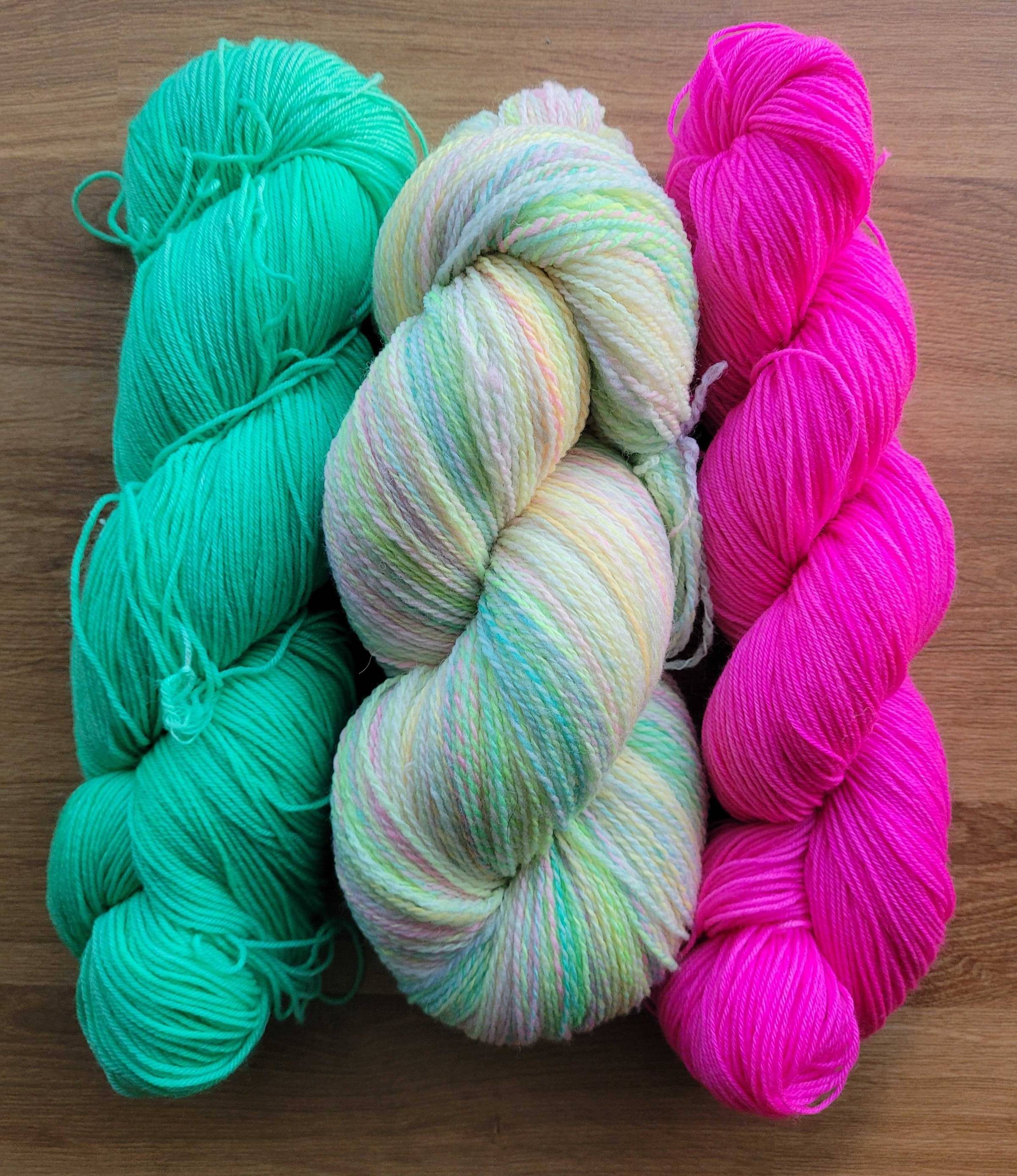 Three bright skeins of yarn. Left to right: neon seafoam green, multicolored neon/pastel rainbow handspun, searingly bright hot pink.