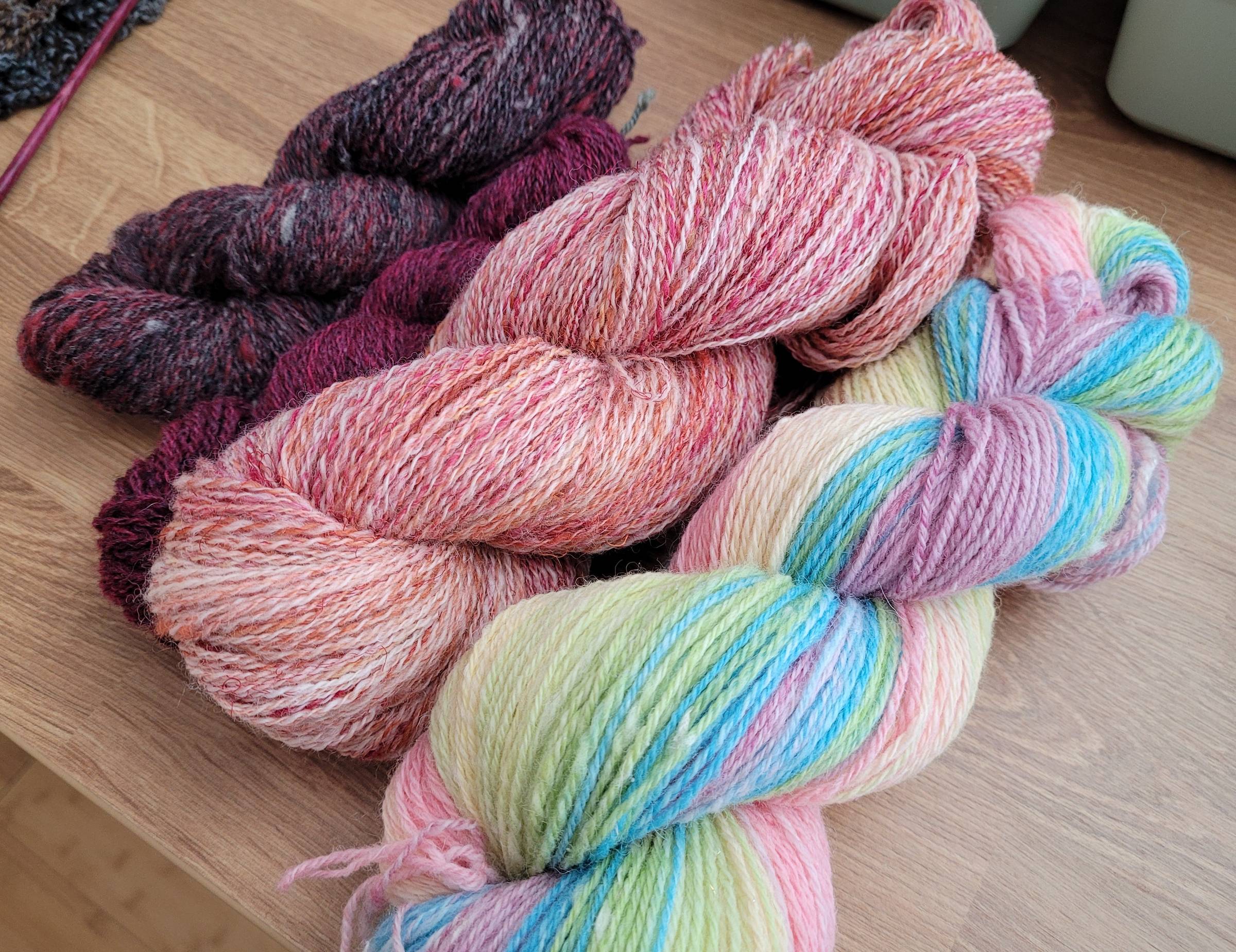 Four handspun skeins of yarn sitting on a wooden work table. Mini skeins are black/gray/red and magenta/black, while the full-size skeins are a two-ply of peachy colorss and white, and a pastel rainbow gradient chain-ply.