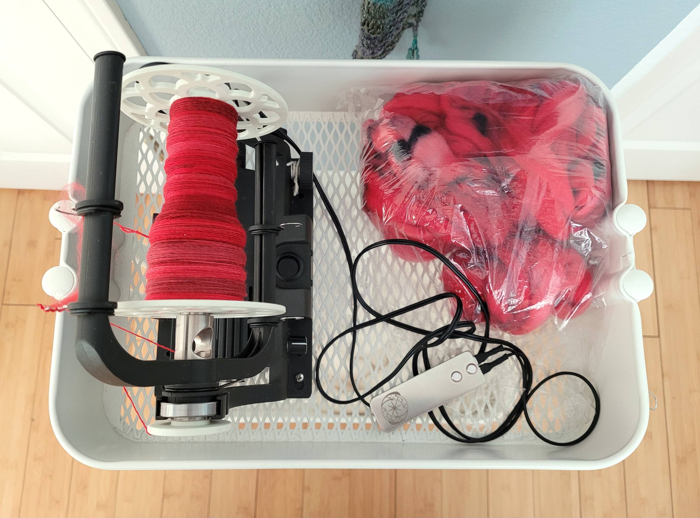 Top-down view of an Electric Eel Wheel (Version 6) with a white bobbin about half filled will tonal red single-ply yarn. A silver control pedal is to the right, alongside a clear bag full of the remaining fiber. All items are sitting in the top tier of a white rolling cart.
