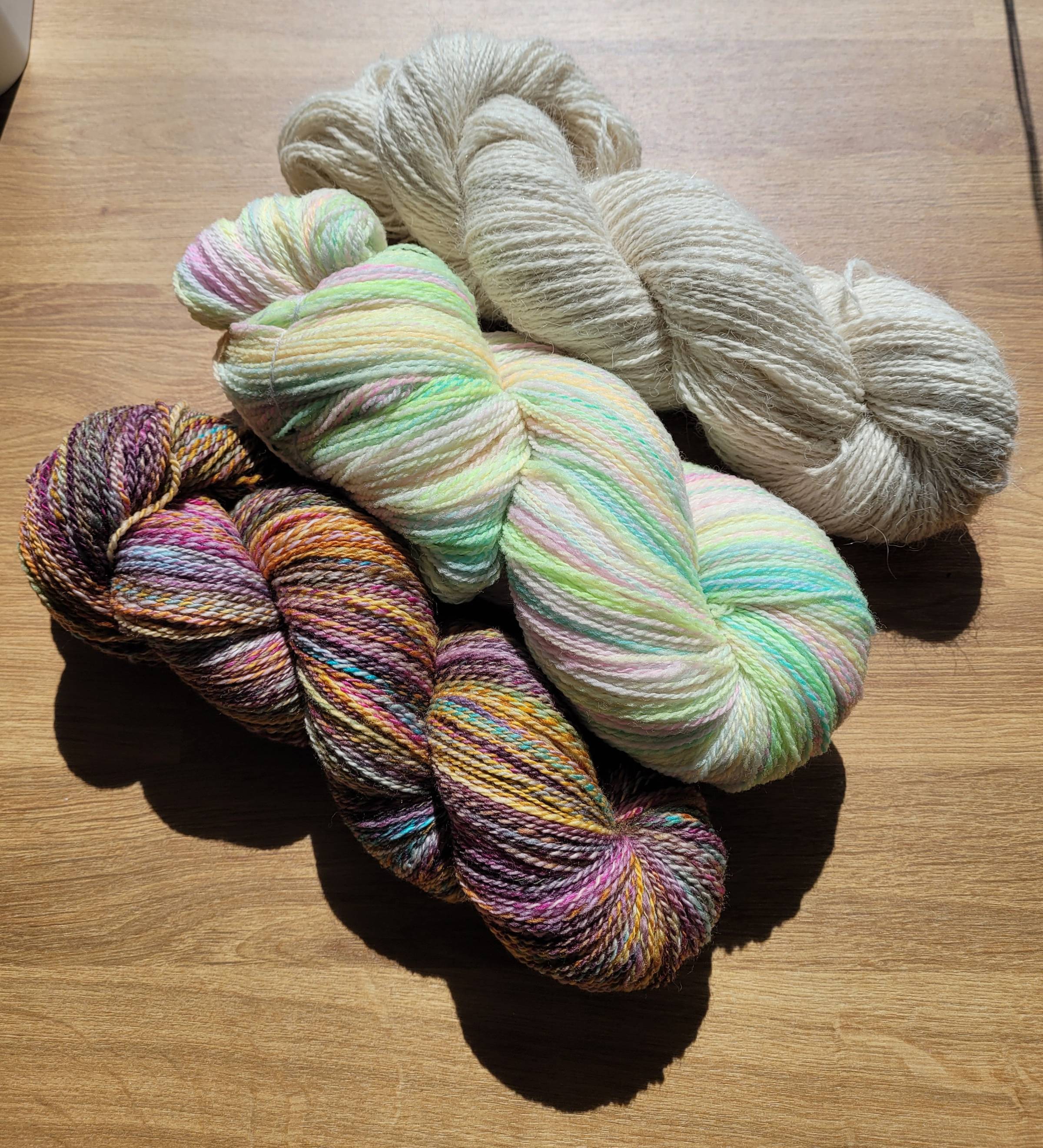 Three skeins of handspun yarn. Bottom: multicolored skein of deep jewel-toned purples, aquas, pinks, and oranges. Middle: Pastel rainbow in a fractal 2-ply. Top: Undyed white alpaca 2-ply.