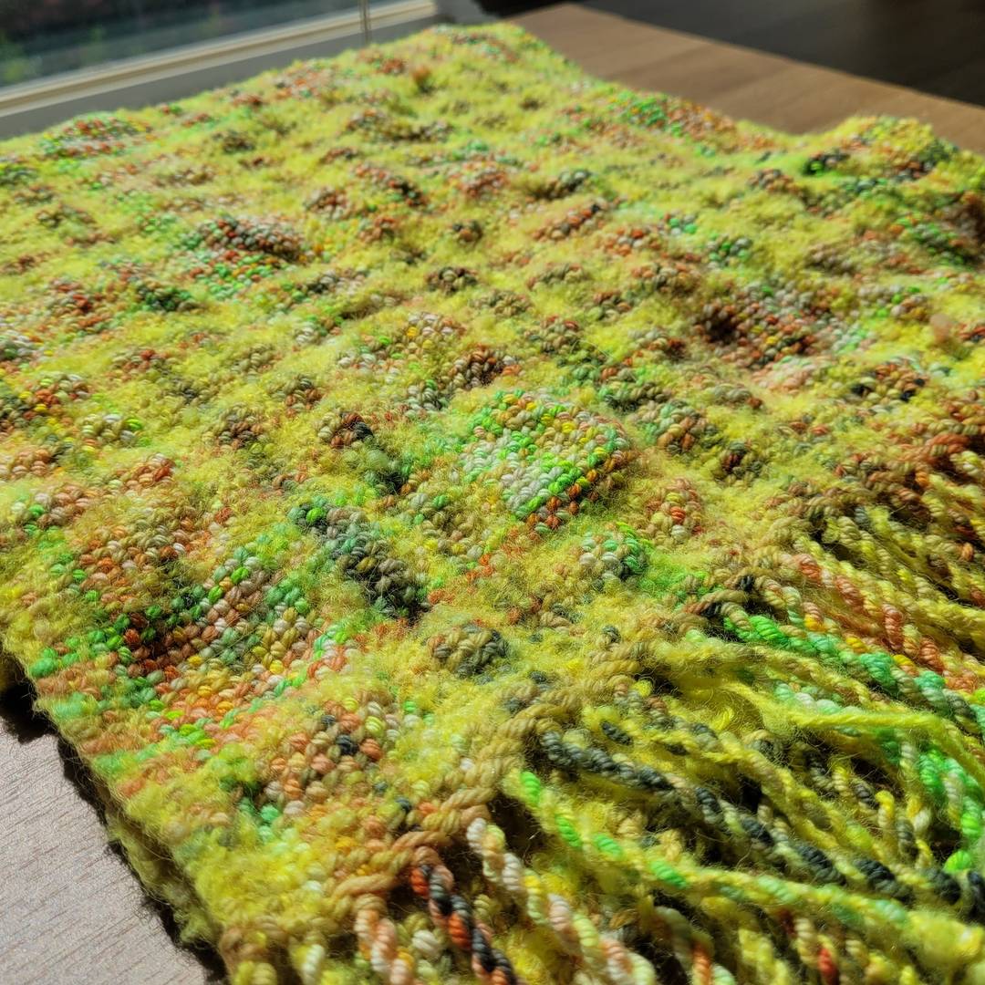 Mostly-yellow plaid scarf with variegated yellow/green/orange blips throughout. Scarf has experienced differential shrinkage, so parts of it are felted and puckered compared to others.
