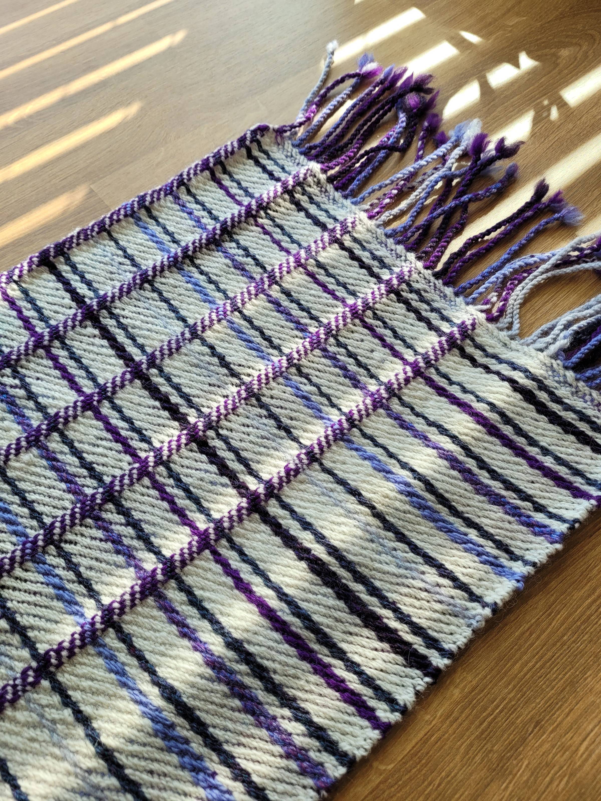 Back side of a handwoven plaid scarf. Visible on this side ais mostly white, with four vertical red-purple stripes, and many small stripes in varying purple shades going horizontally. Fringe is visible at the top.