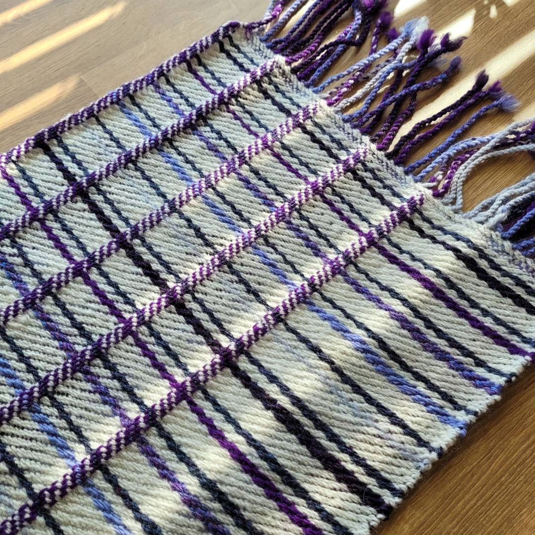 Back side of a handwoven plaid scarf. Visible on this side ais mostly white, with four vertical red-purple stripes, and many small stripes in varying purple shades going horizontally. Fringe is visible at the top.