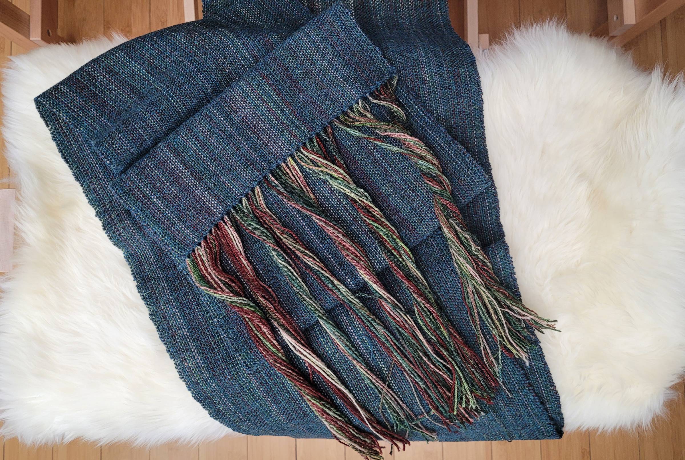 Dark teal handwoven scarf, with brown/green/white earthtones showing in the warp, sitting on top of a sheepskin-covered bench.