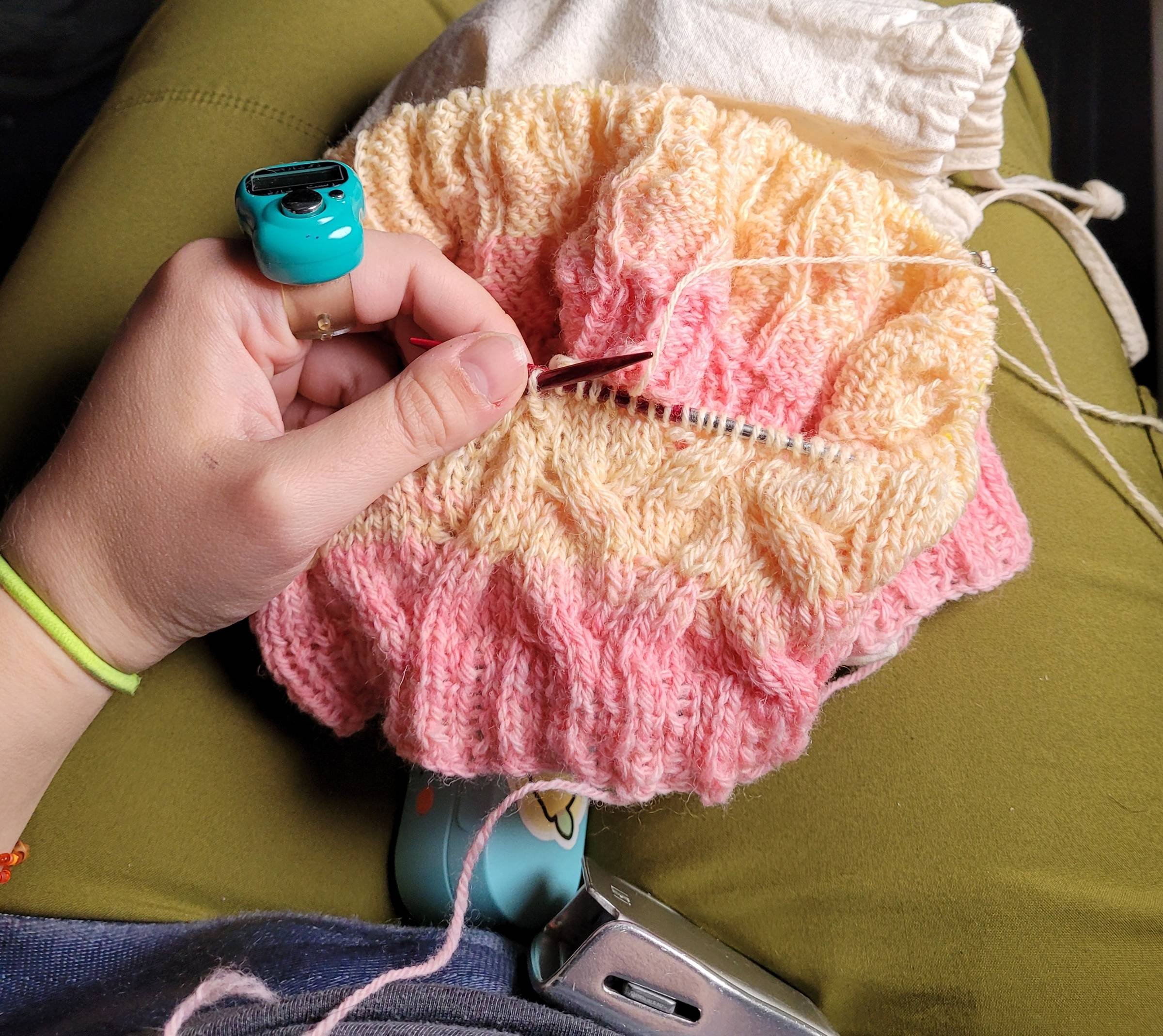 My hand holding the left needle of a knitting project, with a teal row counter on my index finger. Ribbing and cables are visible on the cowl, which started at the ribbing with a peachy pink color and shifted into a very light orange-yellow. An airplane seat belt buckle is visible.