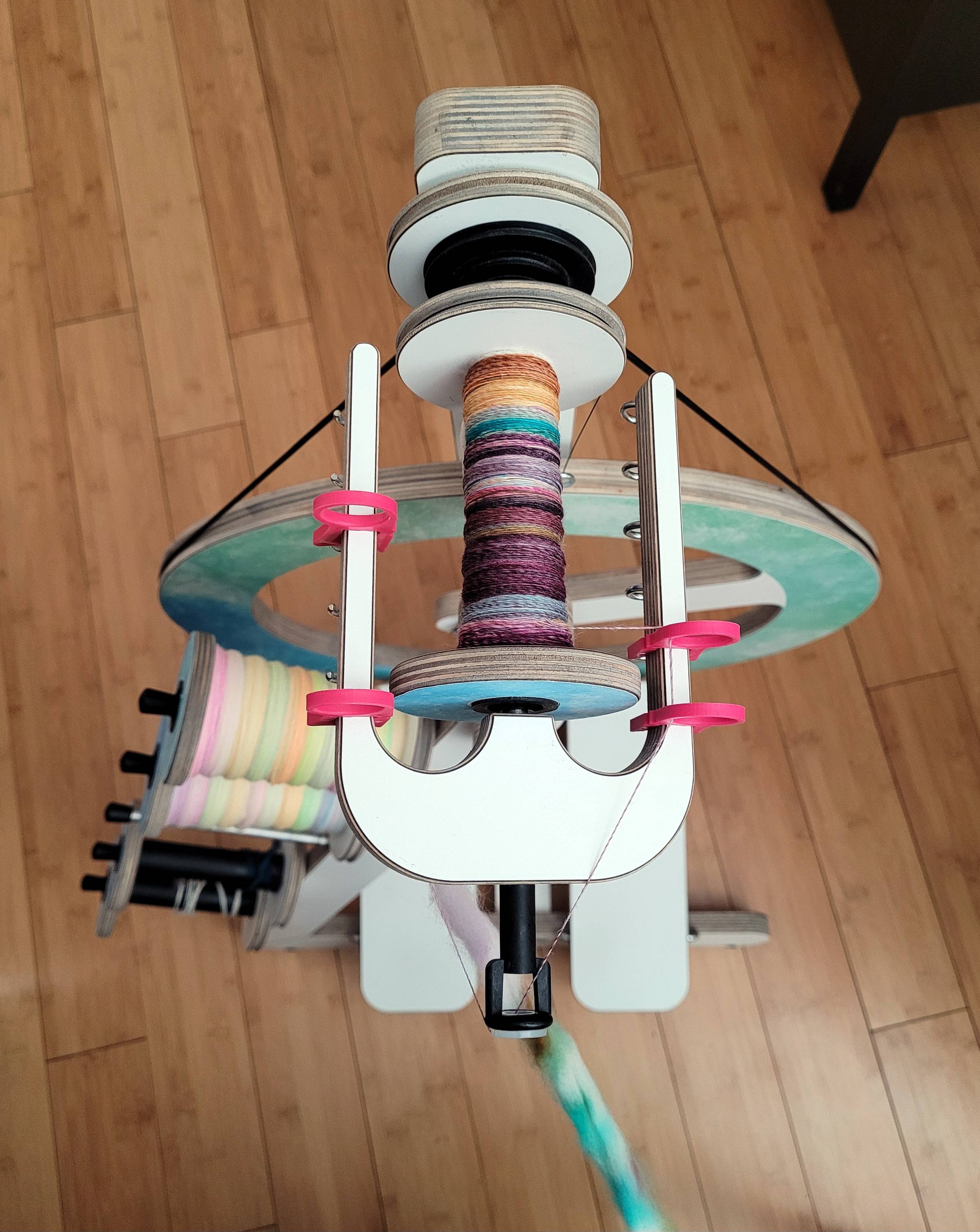 Top-down view of a white Woolmakers Bliss spinning wheel with some watercolor decal modifications. The flyer has magenta, plastic, sliding yarn guides (two on each side), and the yarn on the current bobbin is very multicolored. On the left side, two full bobbins of neon-pastel-rainbow singles are resting.