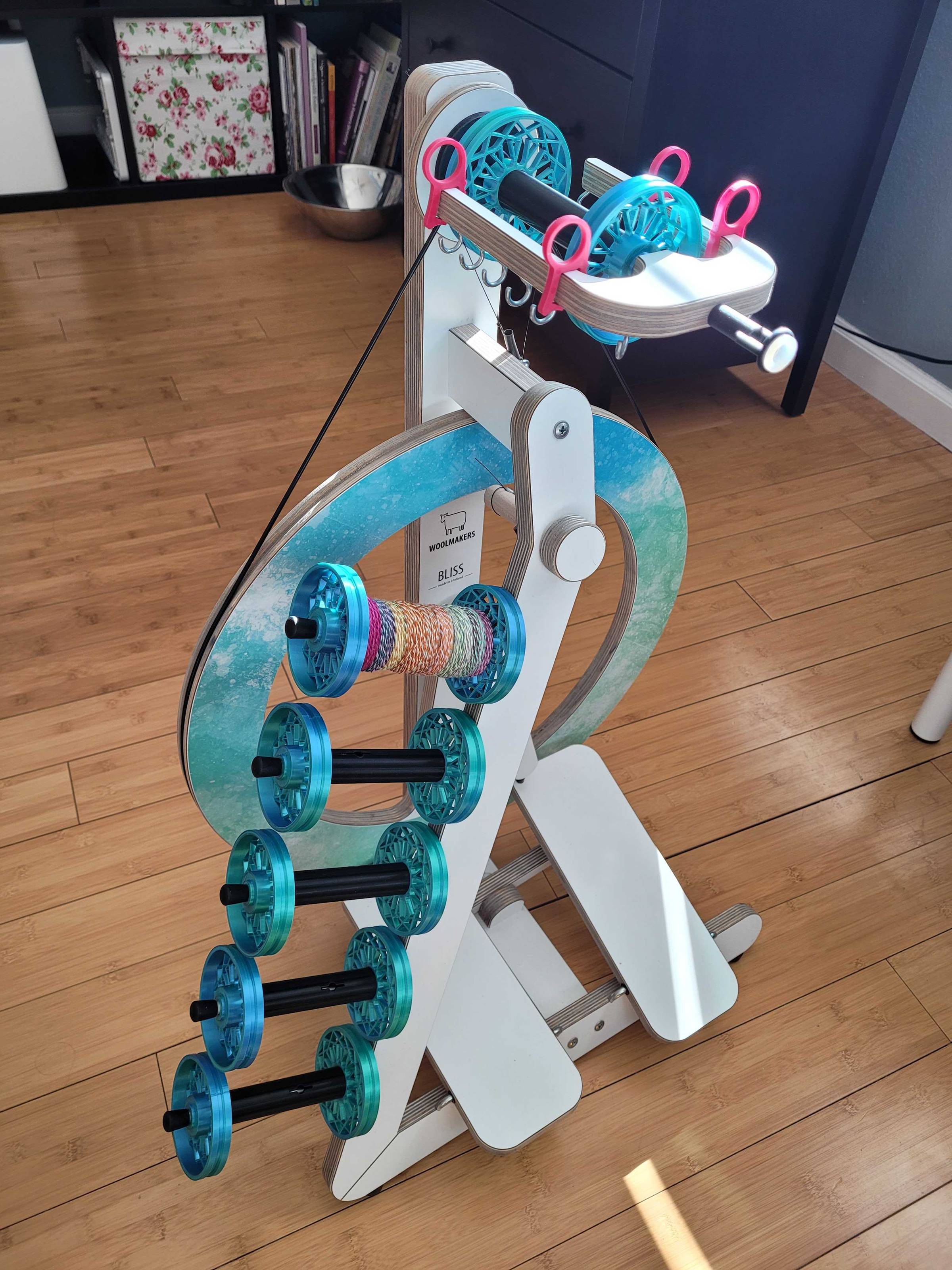 White laminated Woolmakers Bliss spinning wheel, kitted out with shiny blue-green 3D printed bobbins (6) and a custom blue-green watercolor decal on the drive wheel.
