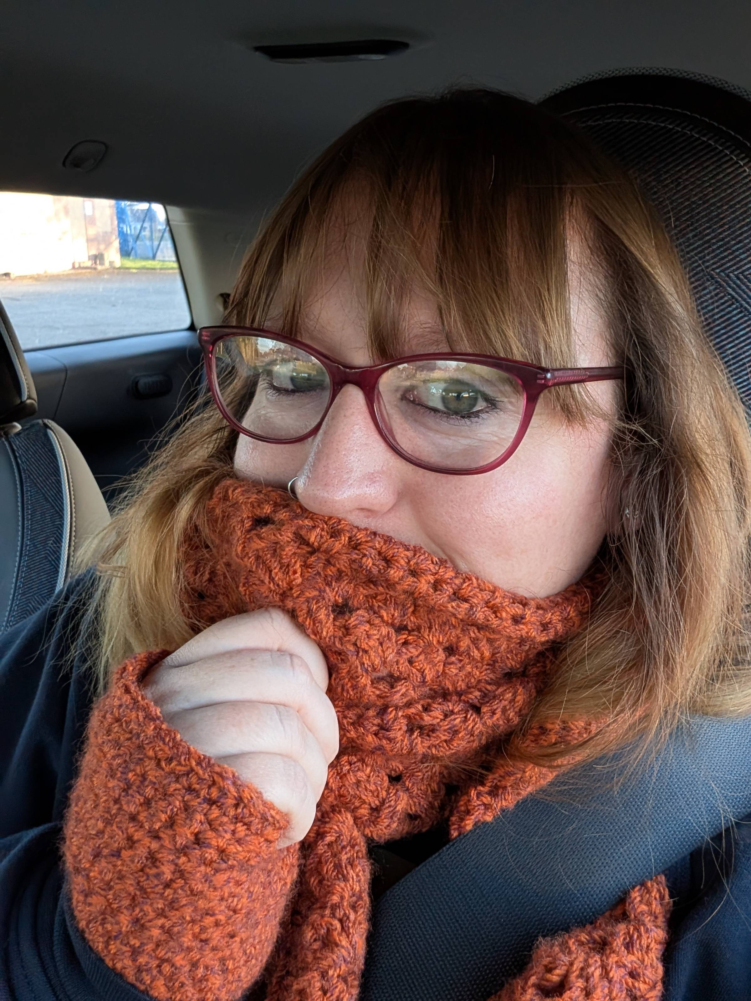 A pale woman with brown hair and glasses cozied in a autumnal orange crocheted scarf and gloves in a car. 