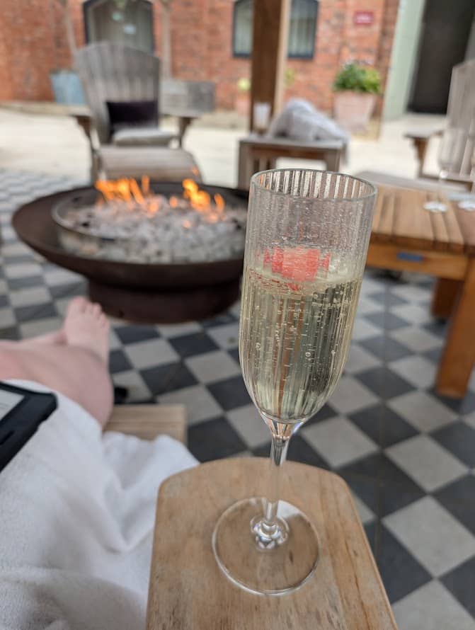A cozy outdoor patio scene featuring a central fire pit surrounded by several wooden chairs. In the foreground, a glass of sparkling wine with a strawberry garnish rests on a wooden armrest or small table. A person sits nearby with bare feet visible, wrap