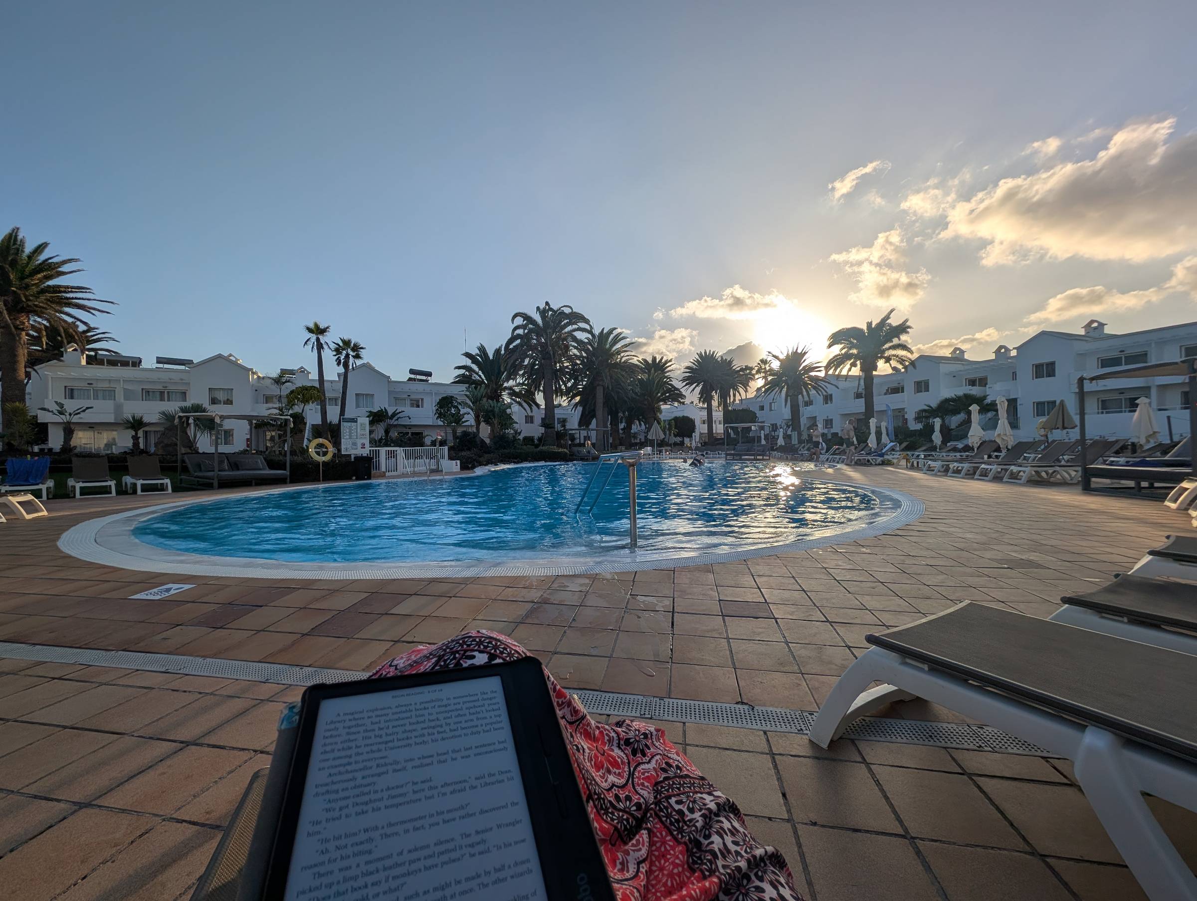 Outdoor resort swimming pool at sunset, surrounded by palm trees and white low-rise buildings. Sun loungers line the tiled pool deck, and the water reflects the warm evening light. A person’s lap with an e-reader is visible in the foreground.