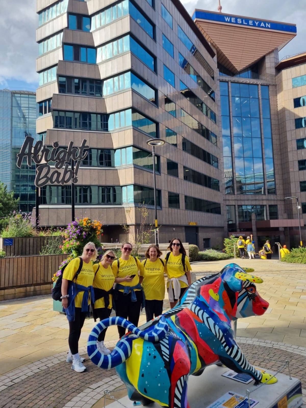 ix people wearing matching yellow "WESLEYAN" shirts stand in front of a brightly painted bull sculpture in a modern urban plaza. Behind them is a glass office building with "WESLEYAN" signage and a playful "Alright Bab!" sign. The area features flower arr