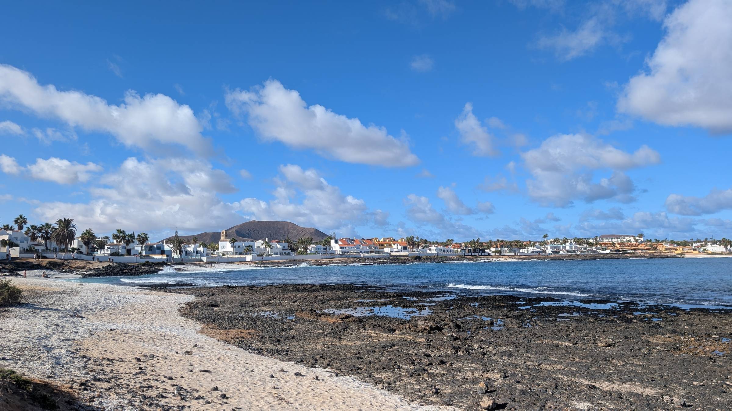 A view of a shoreline of white buildings leading arounf to a harbour. In the background is a, old volcanic crator. In the foreground is a white pebbly beach. The sea is a beautiful blue and the sky is bright with a few clouds.