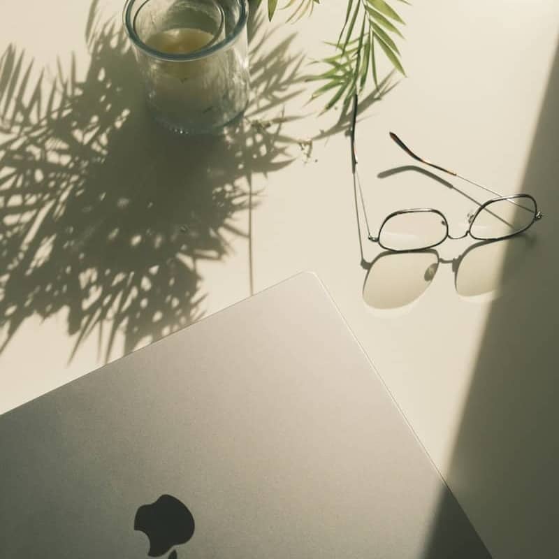 A minimalist scene on a light-colored table featuring a closed laptop with an apple logo, a pair of round glasses resting nearby, a small potted plant casting leafy shadows, and a candle in a glass holder. Sunlight casts soft shadows across the table surface.