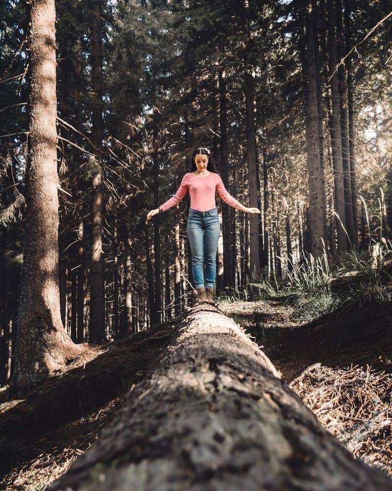 A woman wearing jeans and a pink top walking on a fallen tree trunk, surrounded by trees in the forest and suunlight streaming through the tall, immense treess