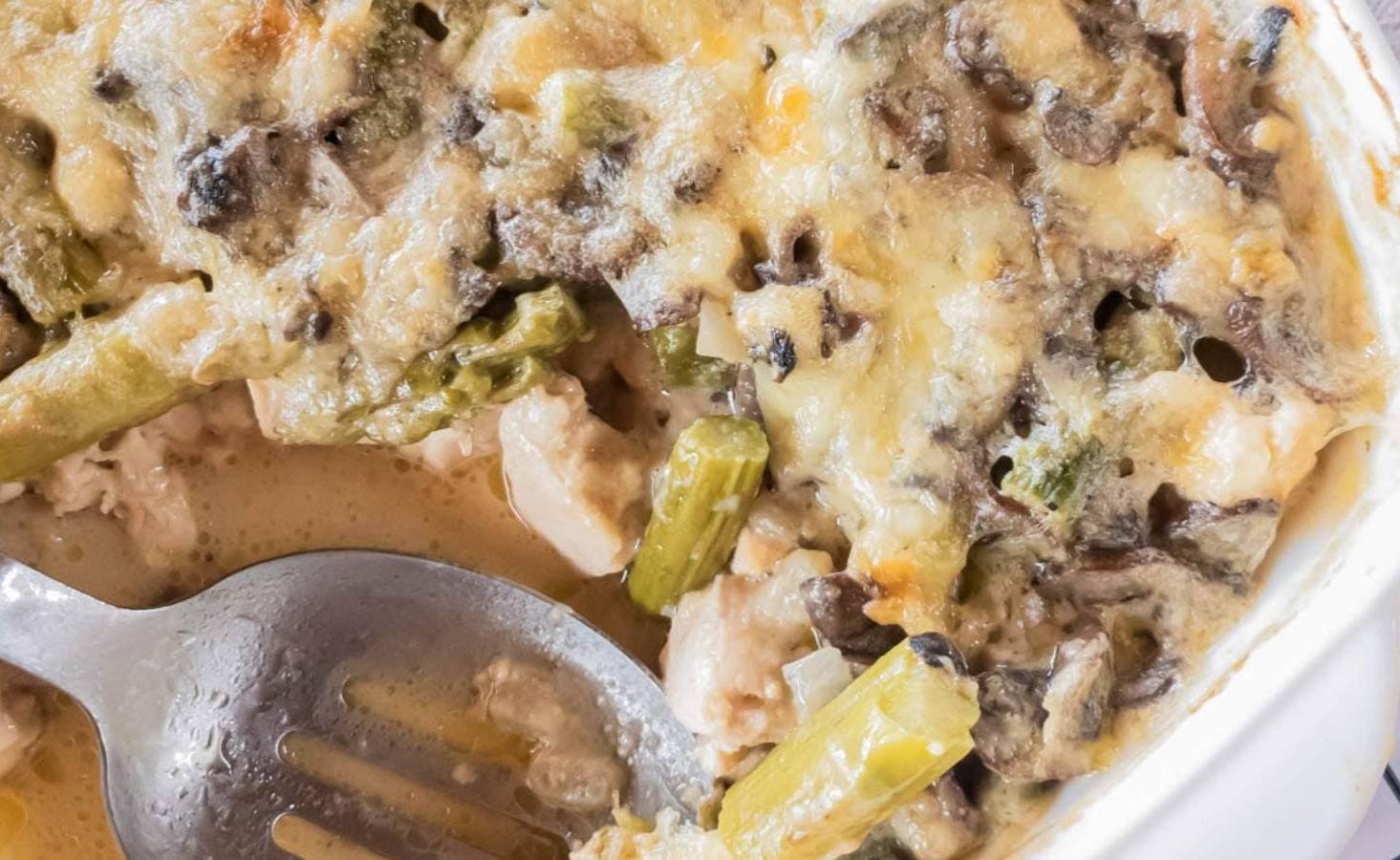 A close-up of a baked casserole with melted cheese, mushrooms, asparagus, and pieces of chicken.