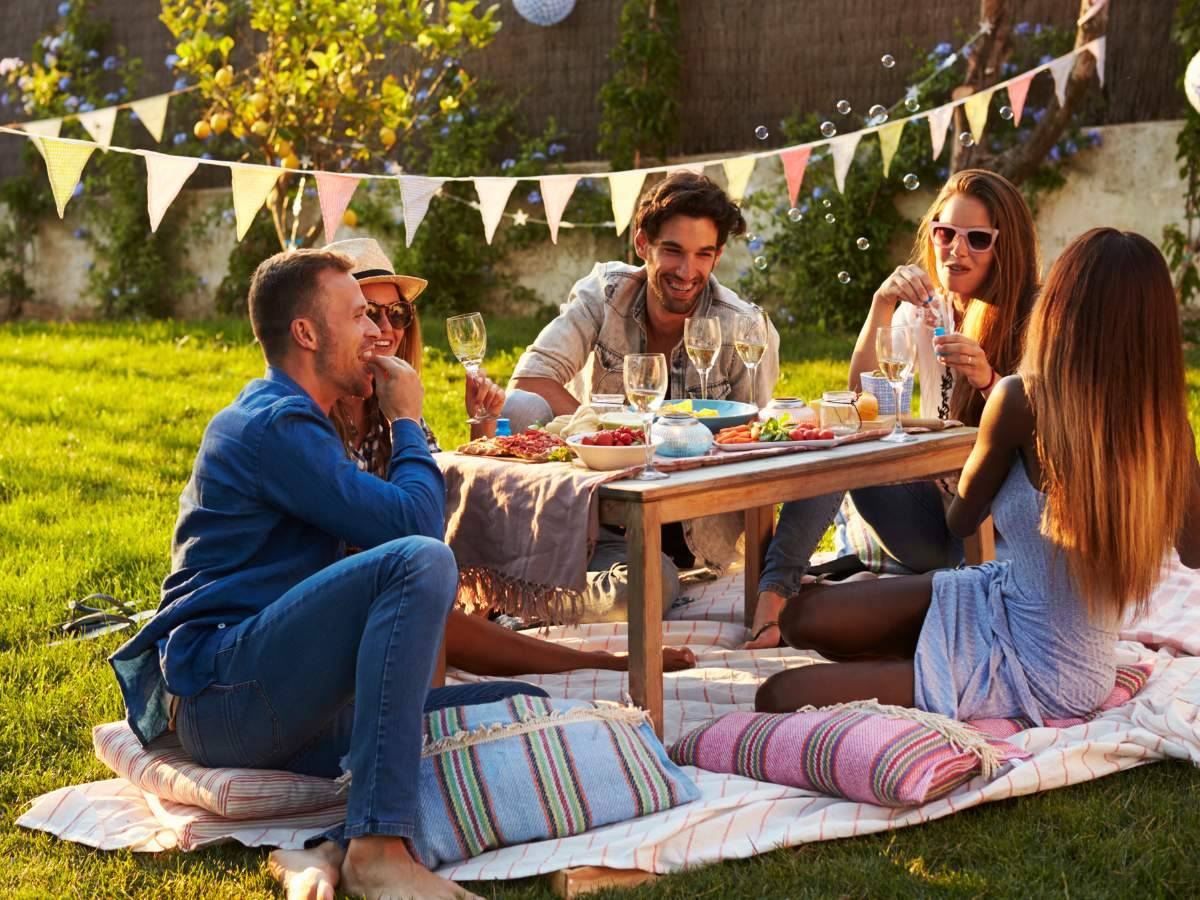 A group of people sitting on a picnic blanket in a garden, enjoying food and drinks.