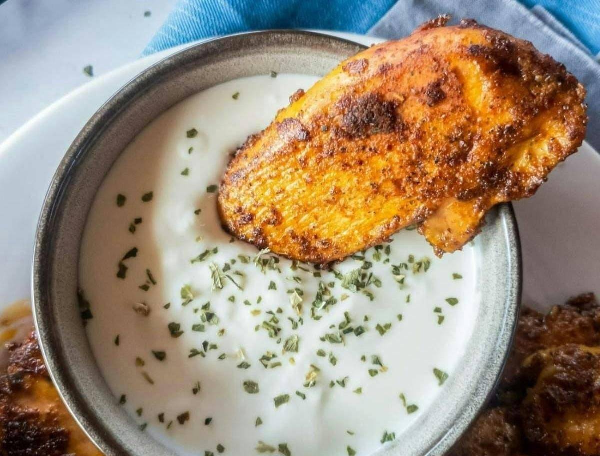 A piece of seasoned chicken being dipped into a bowl of creamy white sauce, garnished with green herbs.