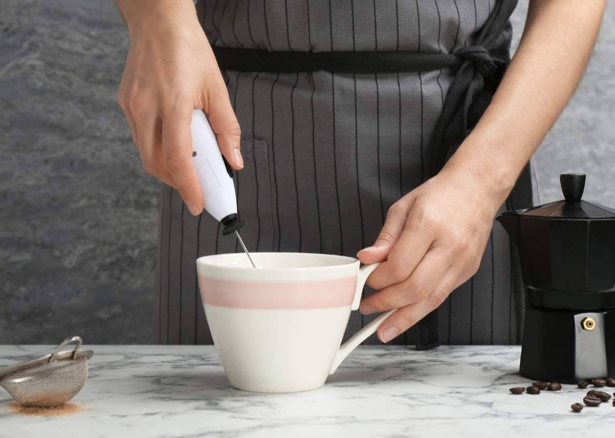 Person using a handheld milk frother in a white mug on a marble countertop. 