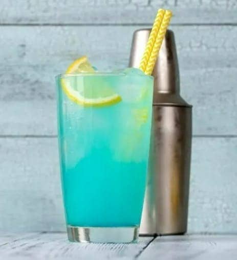 A glass filled with a blue-colored drink, garnished with a lemon slice and a yellow straw, next to a metal cocktail shaker.