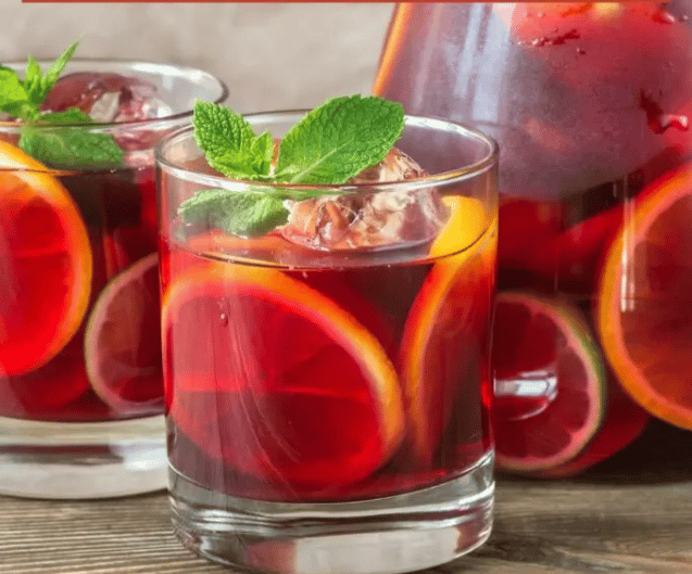 A glass of red beverage with citrus slices and ice cubes, garnished with a mint leaf