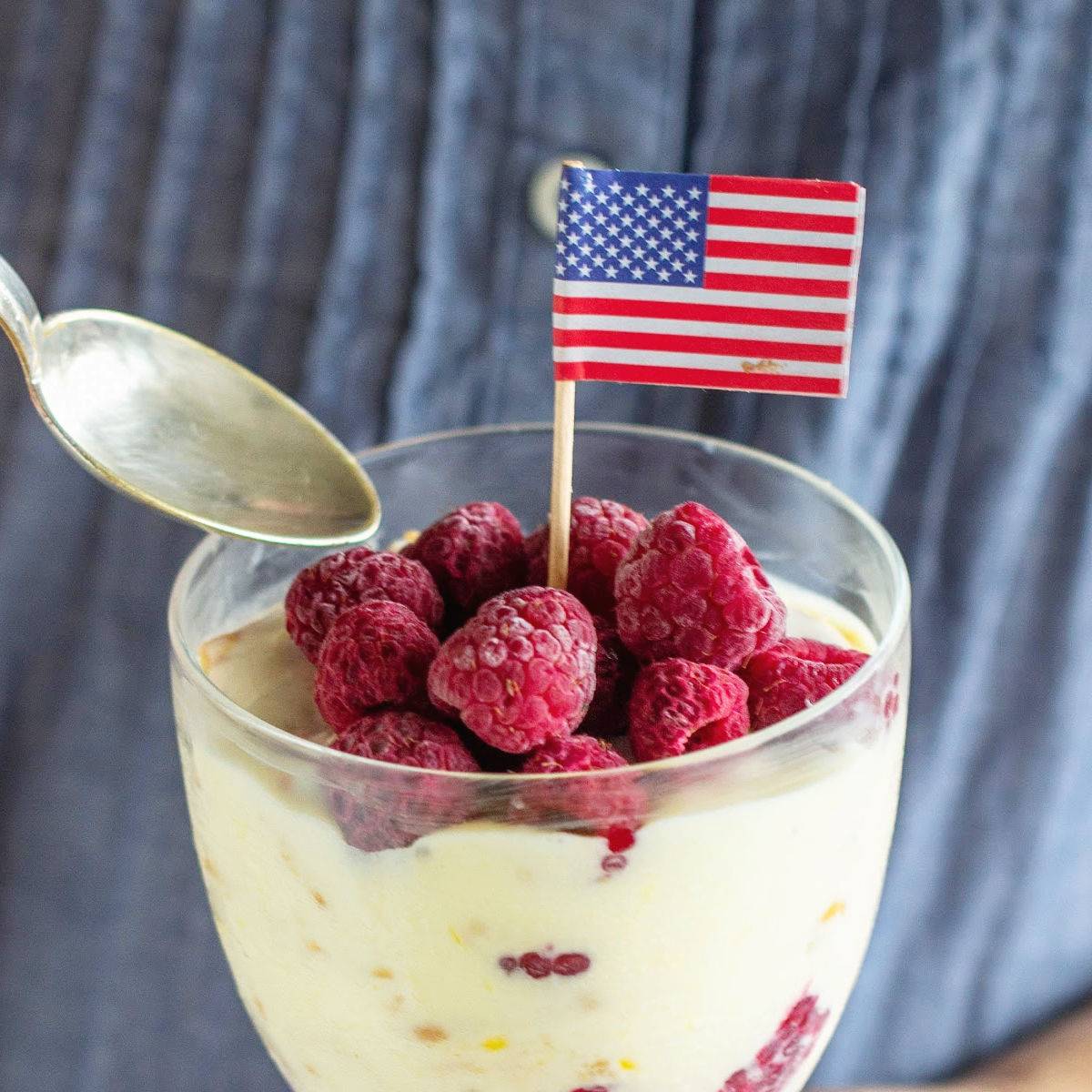 Glass with lemon raspberry dessert and American flag. Silver spoon. Blue background.