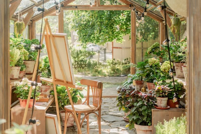 An artist easel and wooden chair inside a sunlit greenhouse filled with potted plants