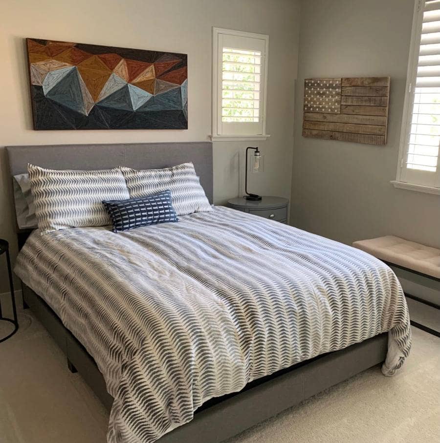 A calm bedroom with a gray upholstered bed, striped bedding, a geometric art piece and plantation shutters