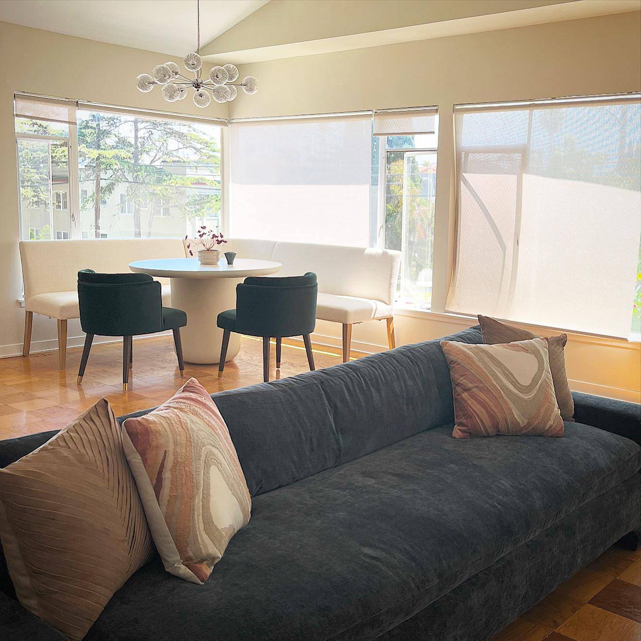 A bright living room with a charcoal sofa and pillows, opening onto a dining nook with a round white table and dark green chairs