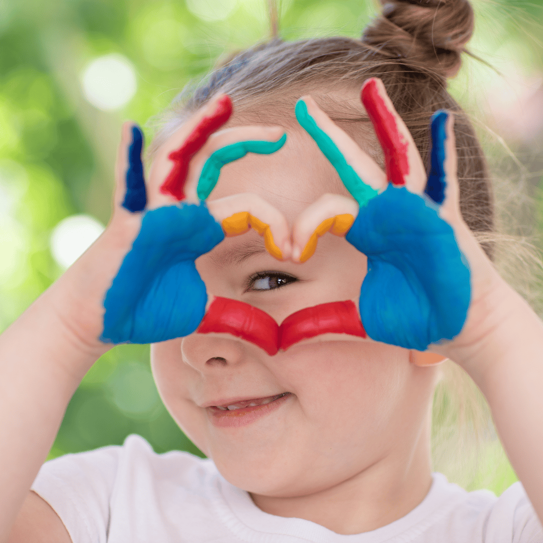 young girl with colorful paint on her hands giving the heart symbol