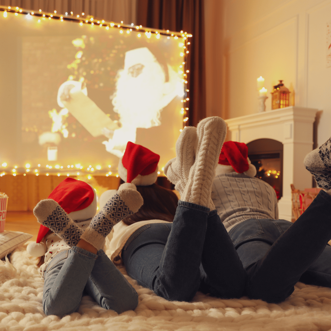 A family in jeans, Christmas sweaters and Santa hats lying on their stomachs facing a screen playing Christmas movies surrounded by twinkling lights