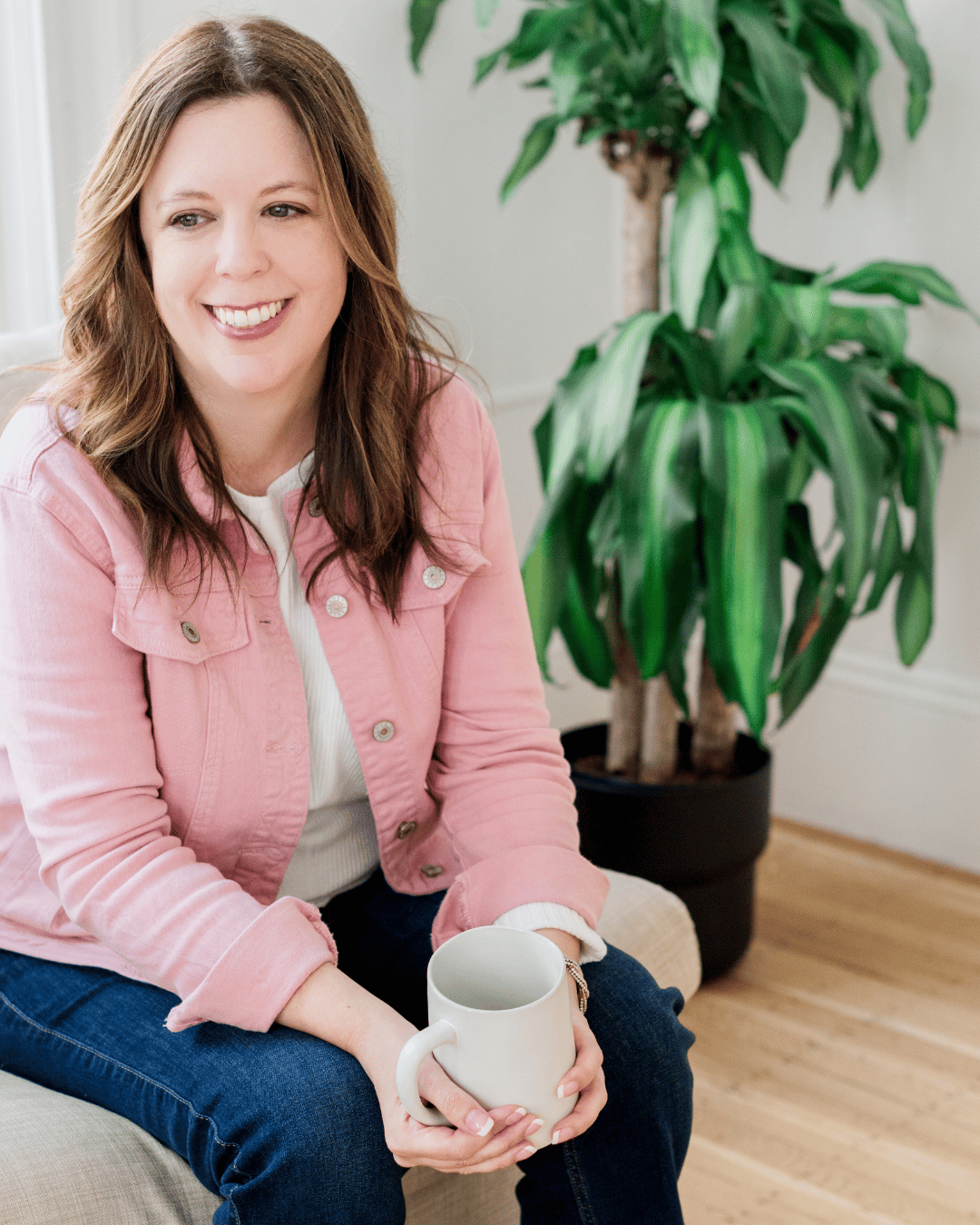 A person with long brown hair is sitting on a chair, wearing a pink jacket and blue jeans. They are smiling and holding a white mug. In the background, there is a large green houseplant.