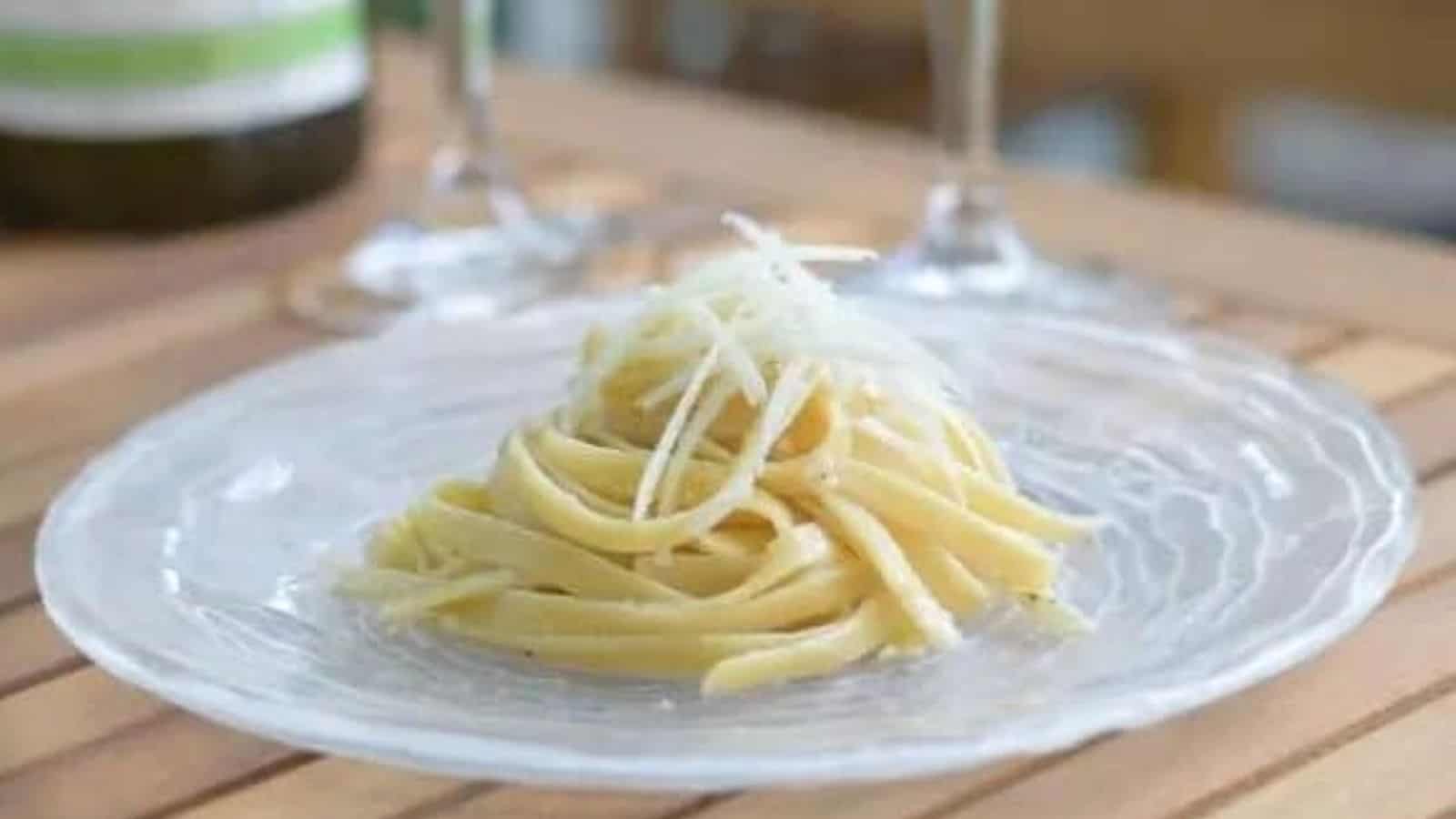 A plate of carefully piled cacio pepe sits on a clear glass plate with wine glasses behind it.