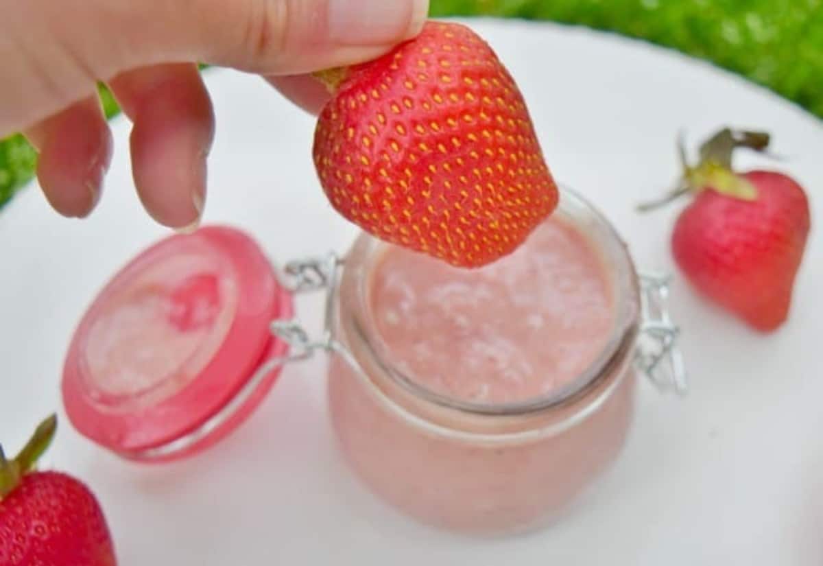 A hand holds a strawberry about to sweep it through a jar of strawberry curd that sits on a white table with more strawberries.