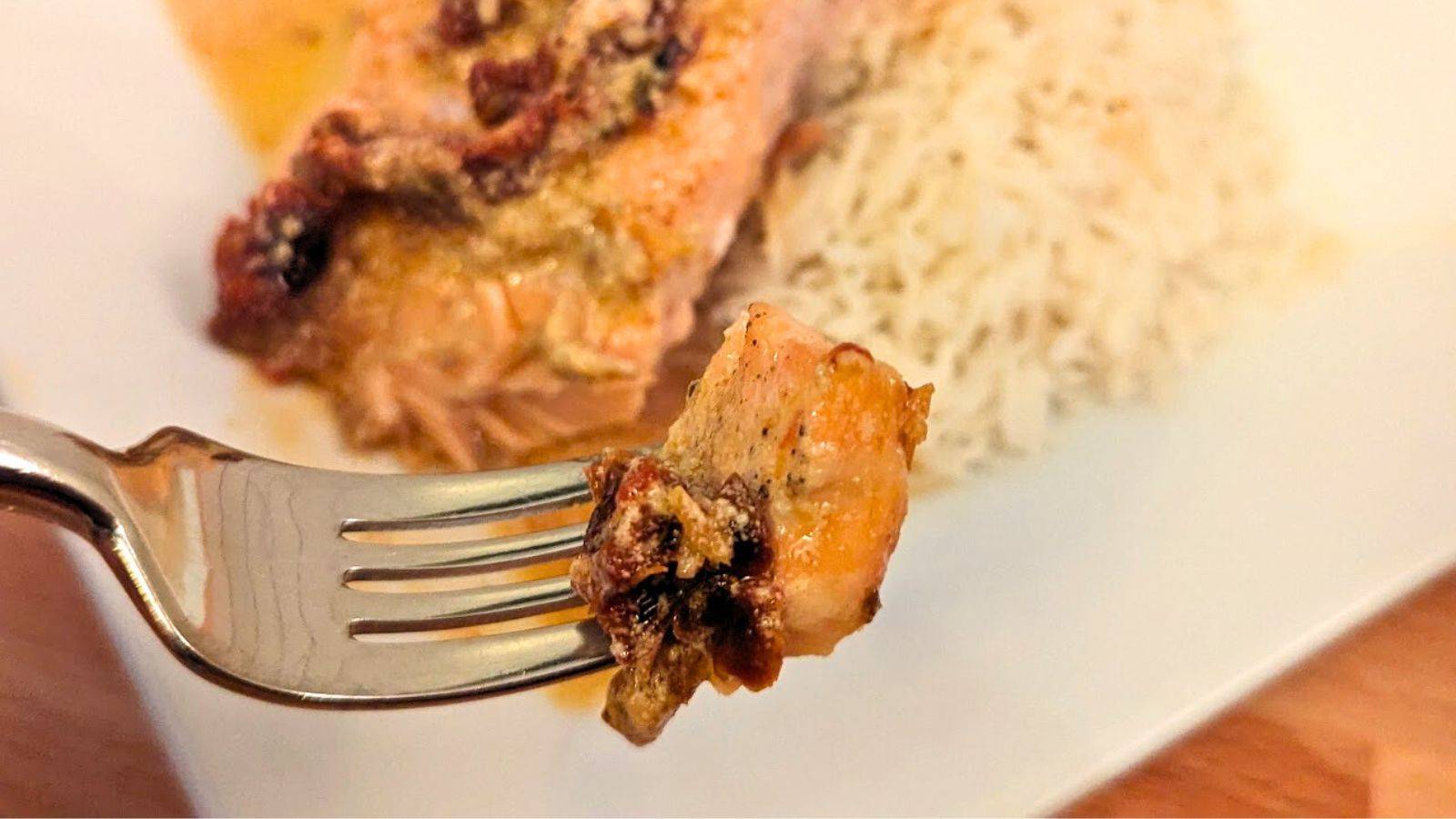 A fork holds a piece of salmon with sun-dried tomato while more of the meal is on a white place with rice in the background.