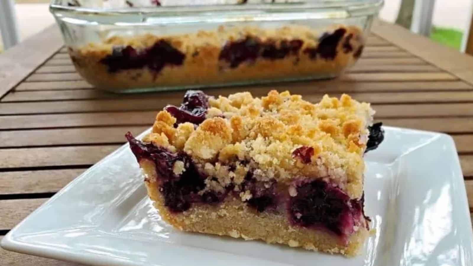 Blueberry crumb bar on a plate in front of a pan with more.