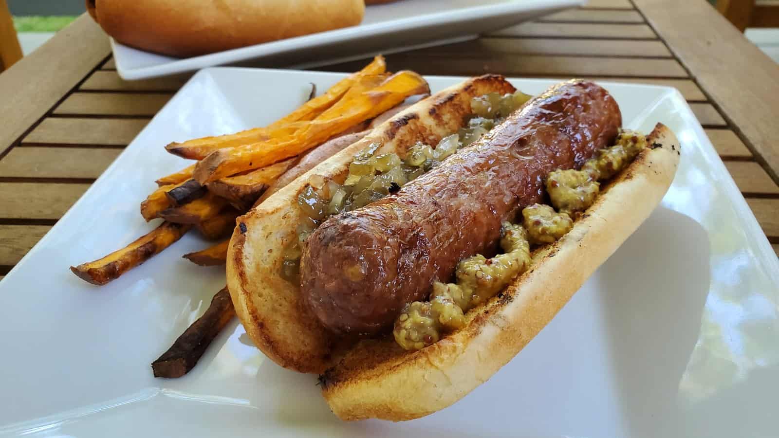 A brat in a bun with mustard on a plate with sweet potato fries.