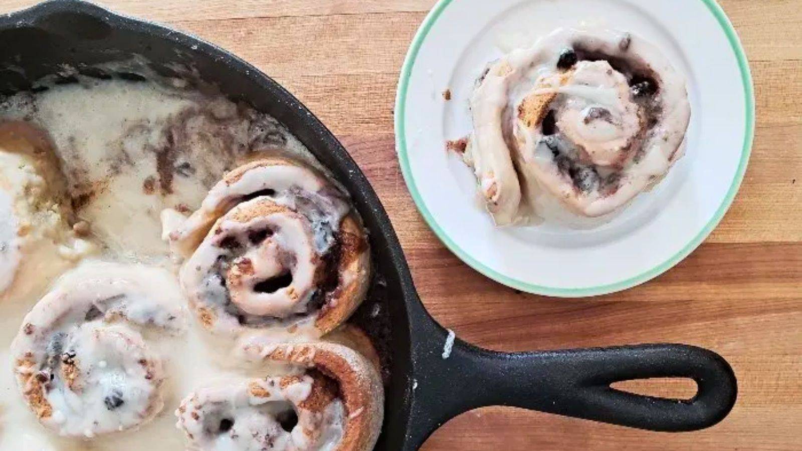 Image shows an overhead of a cast iron skillet with cinnamon rolls and a single one on a plate next to it.