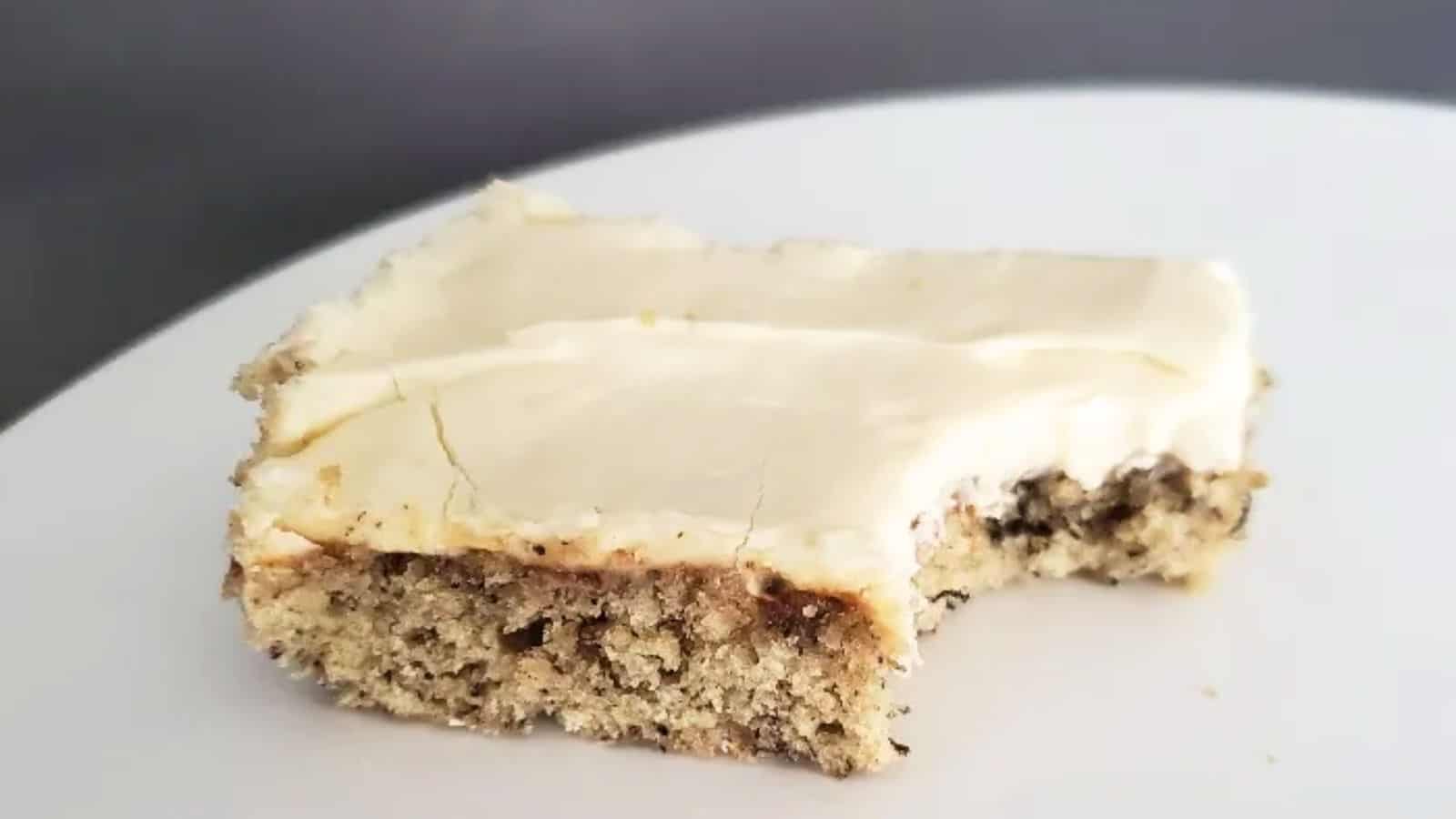 A square of a frosted banana bar sits on a white table with a bite taken out from it.