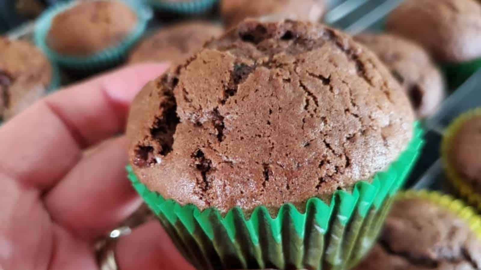 Hand holds a double chocolate muffin in its paper over a batch of more muffins.