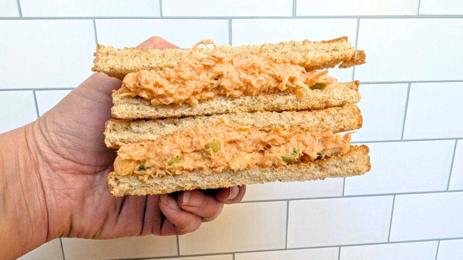 A hand holds two sandwich halves stacked on each other with buffalo chicken salad as the filling.