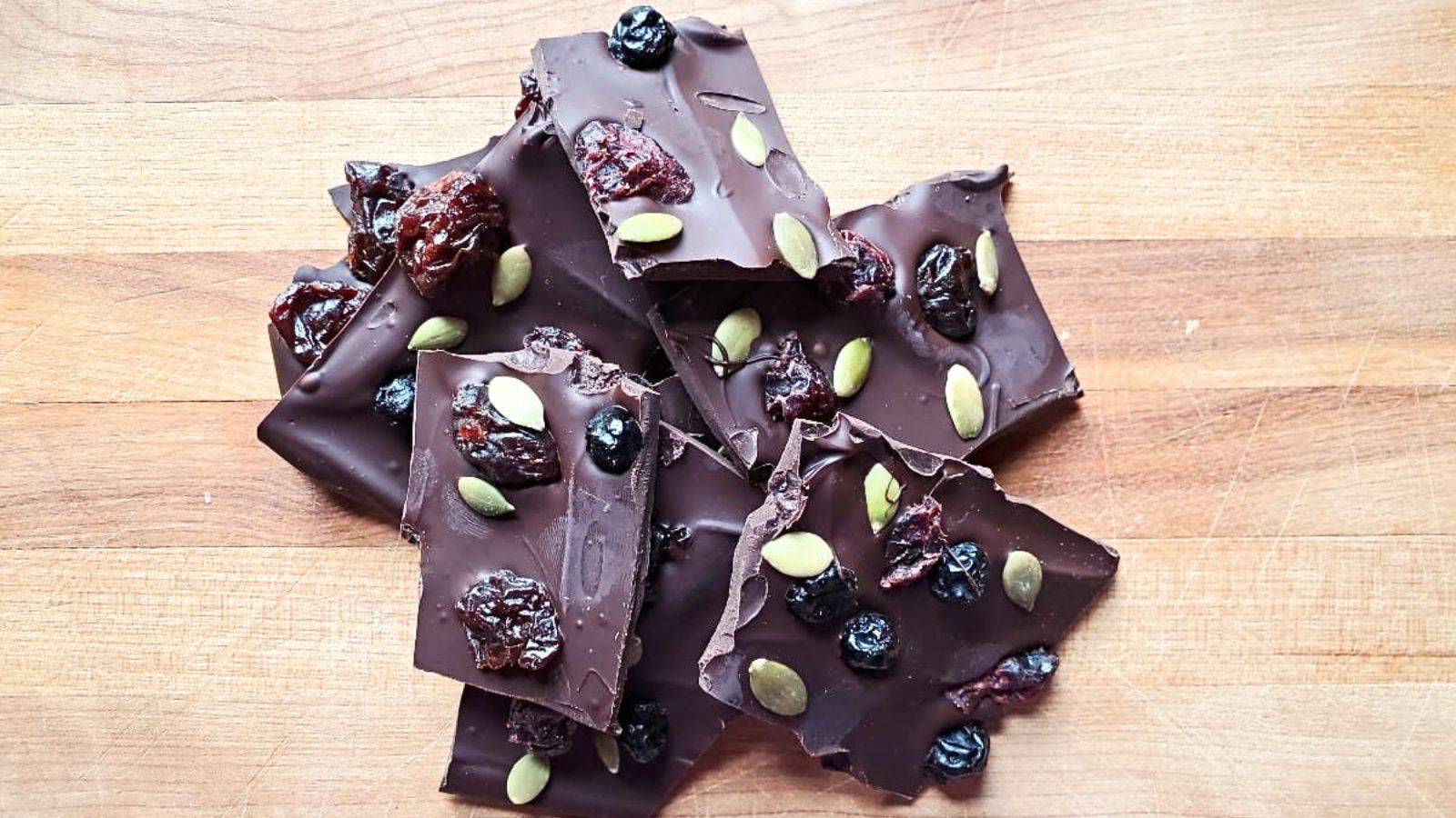 Overhead image of a pile of chocolate bark with dried fruit and seeds mixed in sits on a wooden board.
