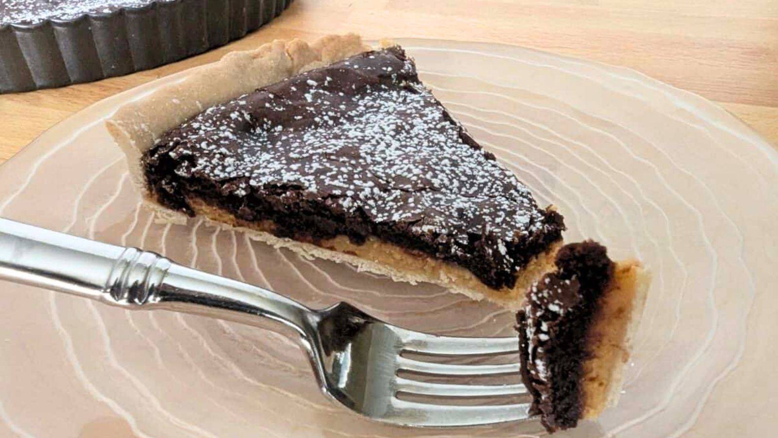 A slice of caramel chocolate tart on a clear round plate with a bite on a fork sitting in front of it.