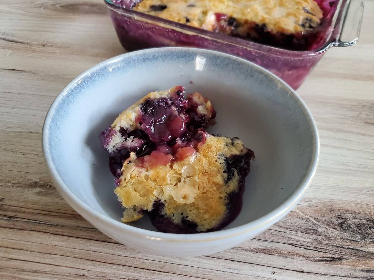 A slice of blueberry pudding cake sits in a blue bowl with the full dish of the batch behind it.