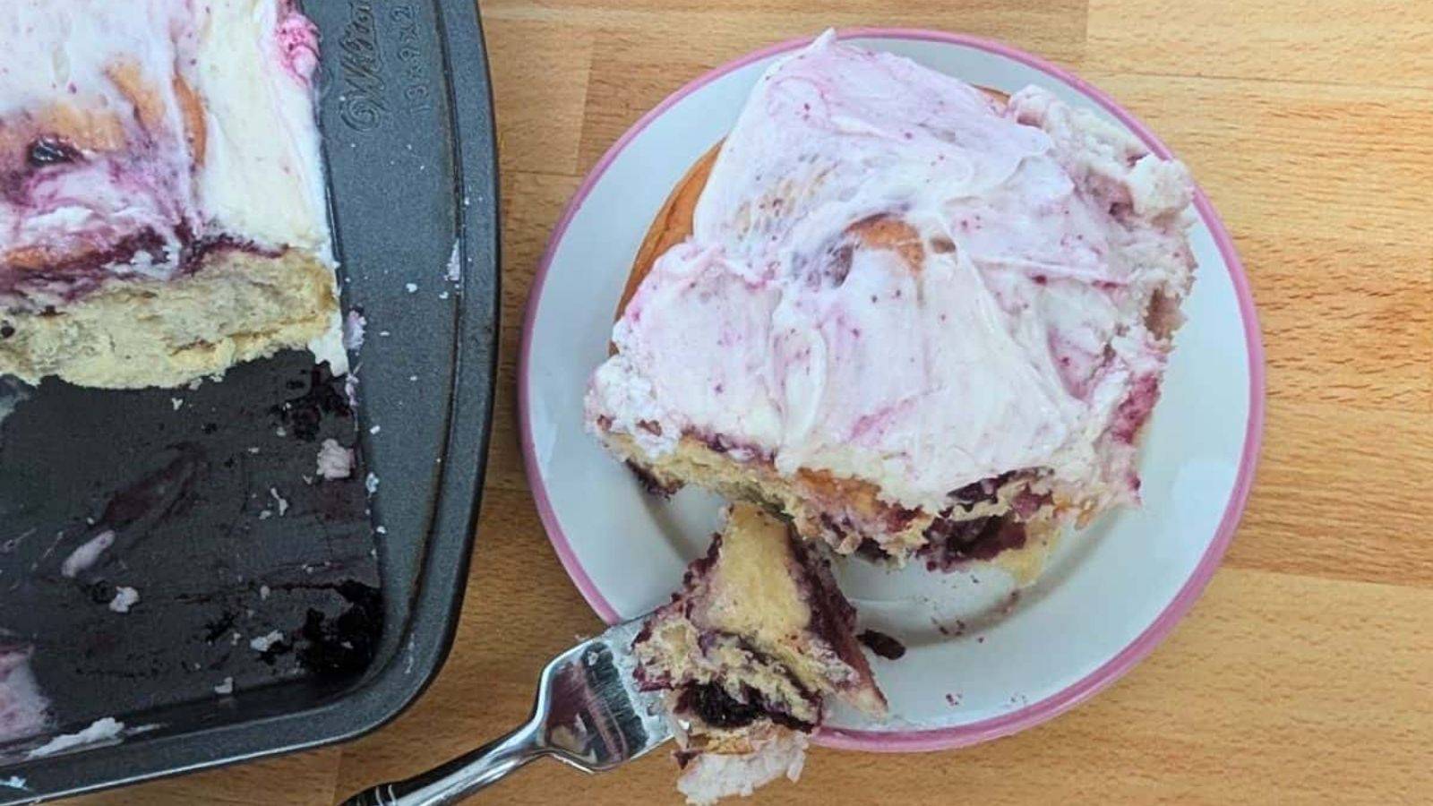 OVerhead of a berry cinnamon roll with a fork holding a bite on a small plate with the pan to the left side.