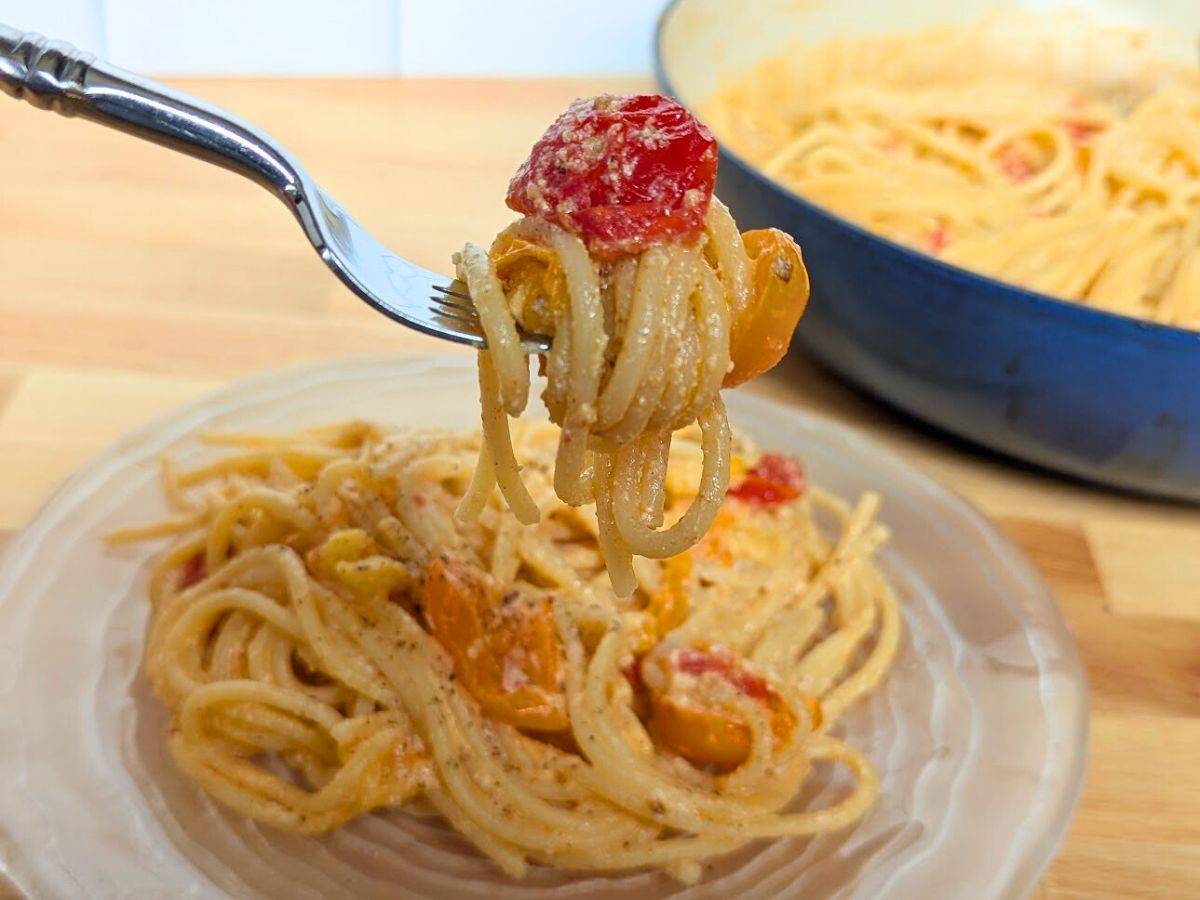 A fork holds a twirl of pasta with a cherry tomato over a plate filled with more pasta and the full pan behind it.